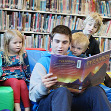 older student reading a book to younger students in a library/