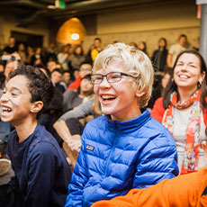 young boy with glasses smiling in an auditorium full of students/