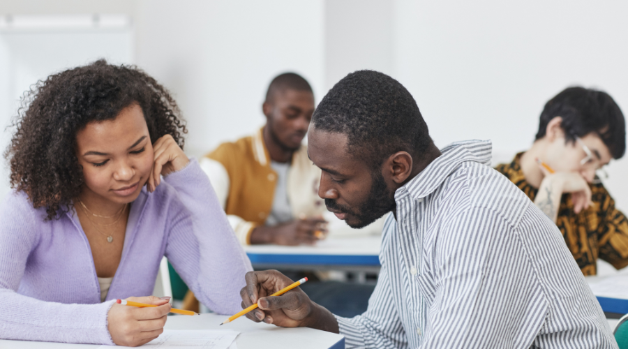 Two African American people reading a paper together in a classroom while two other students look on in the background.