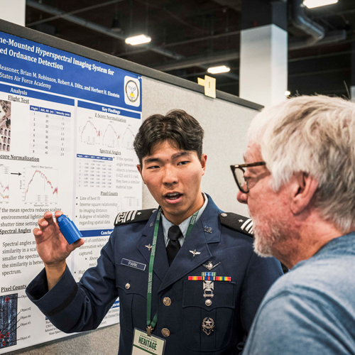 Pacific Islander/Asian-American student in a military uniform presenting a research poster to an older man.
