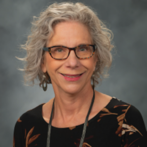 A portrait of Jean Gonsier-Gerdin. She has short, feathered grey hair and glasses and is wearing a black blouse with a brown leaf print.