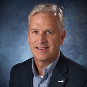 A portrait of Michael Brogioli. He has high silver hair parted to one side. He is wearing an open collared shirt and a black blazer. The background is a blue speckled photographer's backdrop.