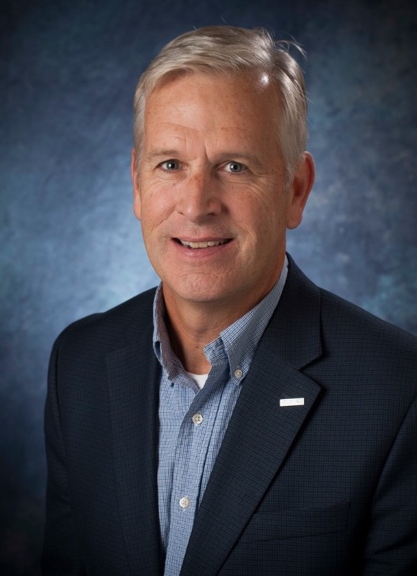 A portrait of Michael Brogioli. He has high grey hair, parted to his left. He is wearing an open collared shirt and a dark suite. He is against a blue speckled photographer's backdrop.