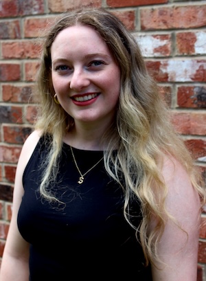 Caucasian woman with long blond hair wearing a black tank top and a gold necklace and earrings in front of a brick background;