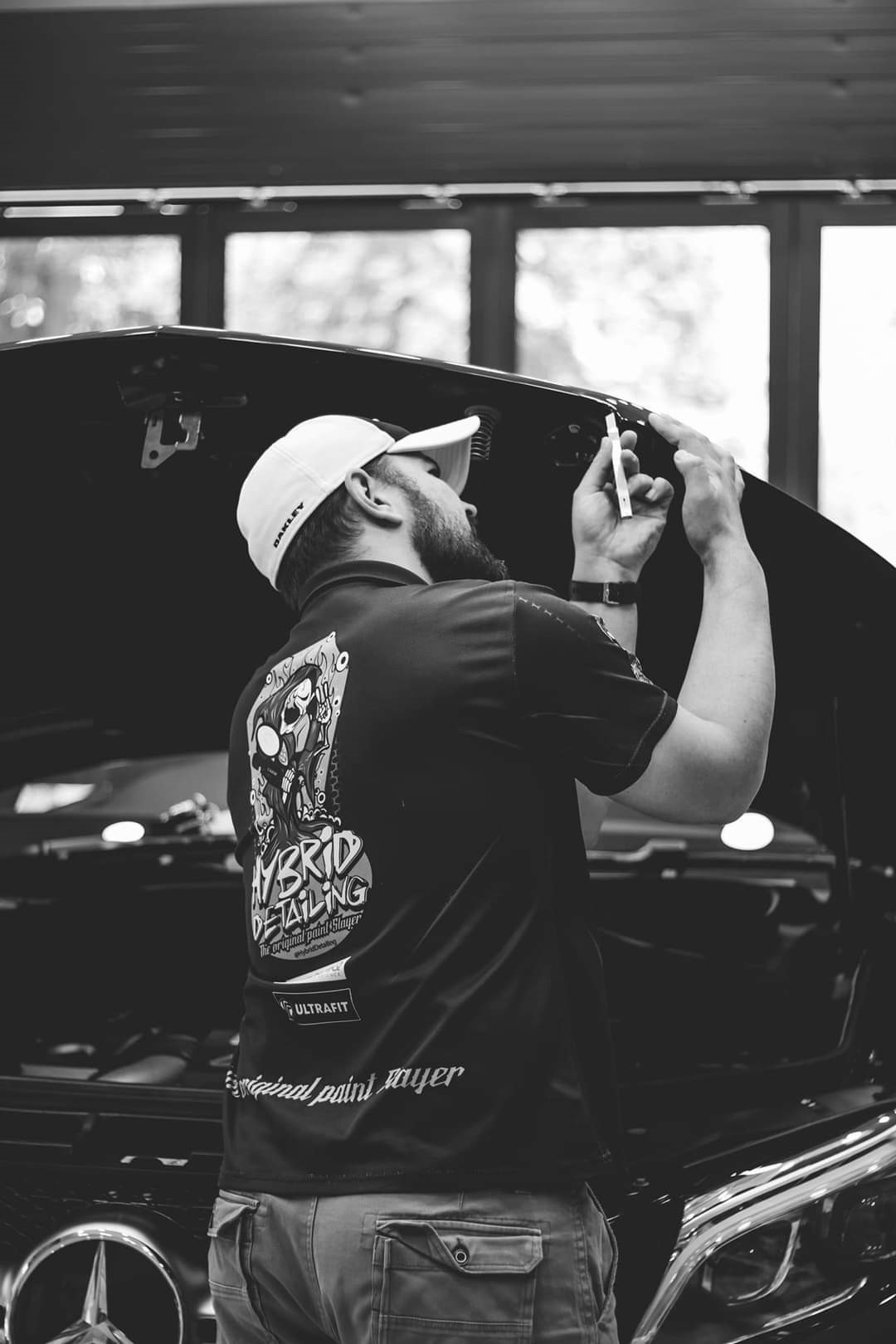 Black and white photo of Hugo van Dyke from the back, wearing a company branded t-shirt, as he stands in front of a car with the hood raised, working on the hood.