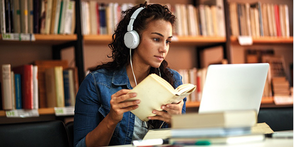 stock photo of a girl in a library using a laptop and holding a book, wearing headphones